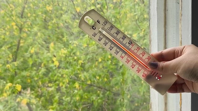 Woman holding a thermometer near a window with yellowing leaves visible outside, representing colder weather, low indoor and outdoor temperatures, autumn and wind.