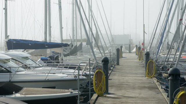Moored yachts line a wooden marina dock walkway with service hoses in dense fog. Harbor pier infrastructure provides safe utility connections and boat berthing during quiet morning weather.