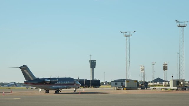 Private business jet stands on the airport tarmac near the air traffic control tower. Corporate luxury aircraft parked on runway apron. VIP executive charter flight and aviation travel concept.