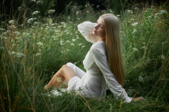 A woman relaxes in a green field filled with tall grass and wildflowers. She poses with long hair flowing as sunlight shines down on her. The scene captures a bright and warm day outdoors