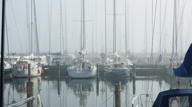 Sailboats moor at wooden marina pilings in dense morning fog. Calm water reflects tall yacht masts and hulls in quiet harbor. Still misty scenery suits nautical travel and maritime boat safety.