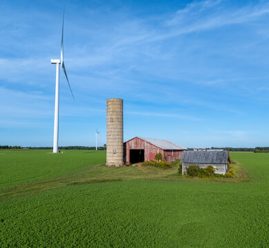 Aerial view of a wind turbine standing next to a rustic barn and a tall silo in a vibrant green field under a clear blue sky in Alma, Michigan, United States.