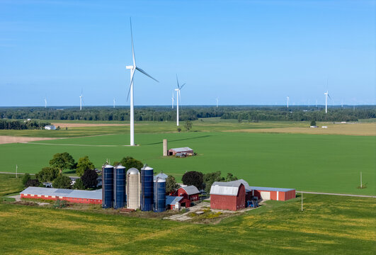 Aerial view of wind turbines towering over a farm with silos and red barns surrounded by green agricultural fields in Alma, Michigan, United States.