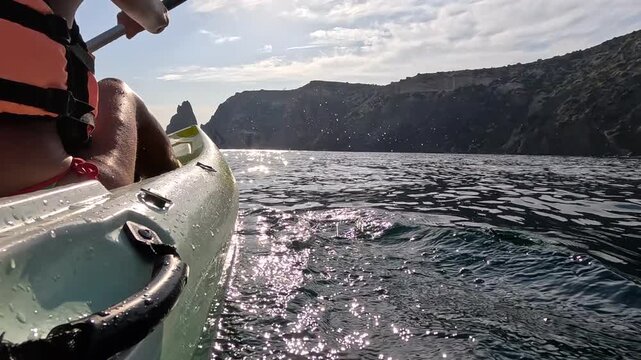 Kayaking an orange paddle through calm ocean waters toward rocky coastal cliffs under a bright sunny sky. Low angle action camera tracking shot.