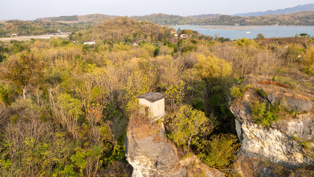 Aerial view of a small stone structure perched on a rocky outcrop surrounded by dense trees and lush vegetation near a calm lake in Klaten, Central Java, Indonesia.