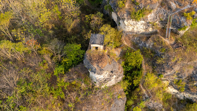 Aerial view of a small stone structure perched on a rocky cliff surrounded by lush trees and vegetation in Klaten, Central Java, Indonesia.