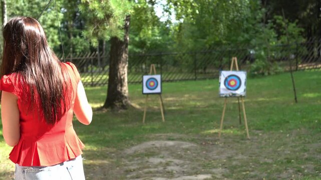 Brunette European girl in vintage jacket and jeans shoots retro crossbow at targets in the park in summer during holiday as an outdoor concept
