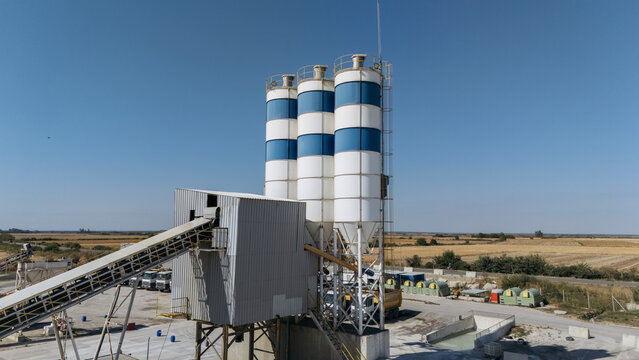 Aerial view of the TASYAPI concrete batching plant featuring three tall white and blue silos, a conveyor belt system, and industrial trucks in a rural setting in Kuzmin, Vojvodina, Serbia.