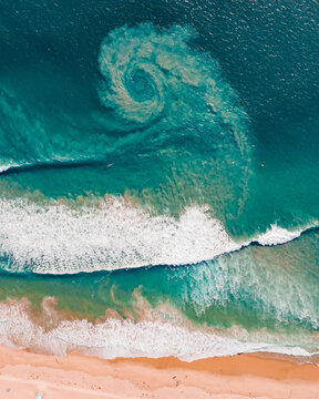 Aerial view of a unique swirling pattern in the turquoise ocean water with white breaking waves hitting the sandy shore in Huntington Beach, California, United States.