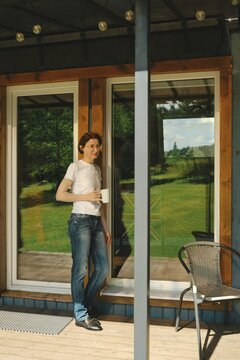 Middle-aged woman leans against wooden frame of large window of wooden cabin while holding cup. Green grass is visible in reflection. Sunlight shines through the window showing bright and open space
