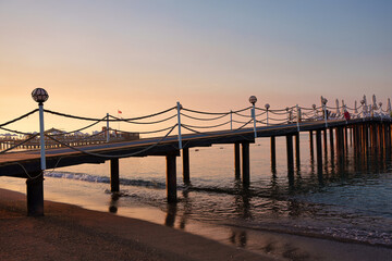 Fototapeta premium Long wooden pier with rope railings on sandy beach during golden sunrise at luxury resort