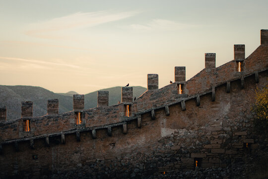 A curving crenellated fortress wall lined with weathered merlons overlooks distant blue mountain ranges beneath a soft cloud-streaked dusk sky, creating a peaceful and atmospheric medieval hilltop sce