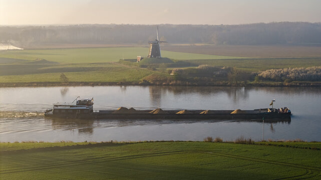 Aerial view of a cargo barge transporting sand along the Meuse river, passing a traditional Dutch windmill in the rural landscape of Beesel, Limburg, Netherlands.