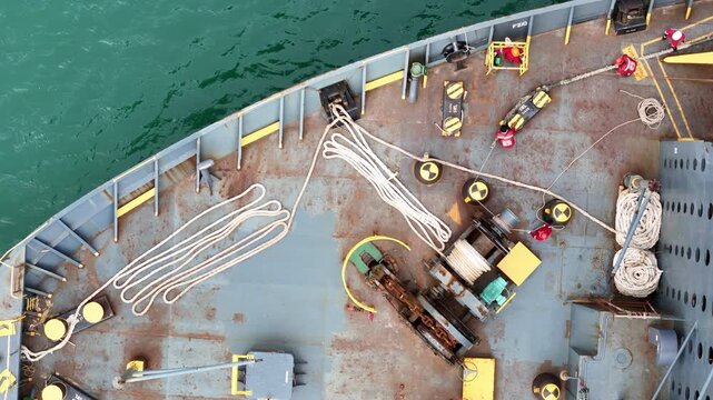multiple ship crew members in bright safety orange working with massive ropes and heavy machinery on the vibrant green deck of a large vessel's bow, with blue water surrounding