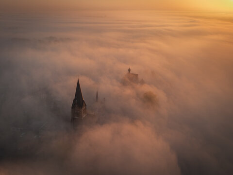 Aerial view of a church spire emerging from thick morning fog at sunrise in Kessel, Limburg, Netherlands.