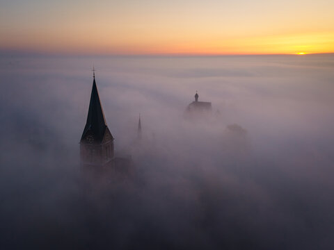 Aerial view of a church spire and historic building emerging from thick morning fog at sunrise in Kessel, Limburg, Netherlands.