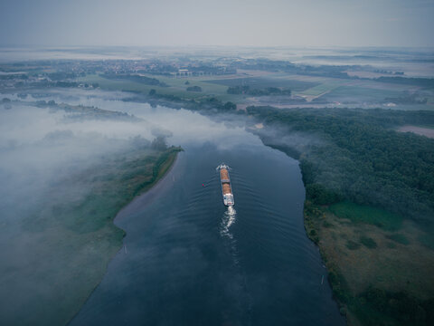 Aerial view of a cargo ship navigating a calm river through misty fields and lush green forests in the early morning light in Kessel, Limburg, Netherlands.