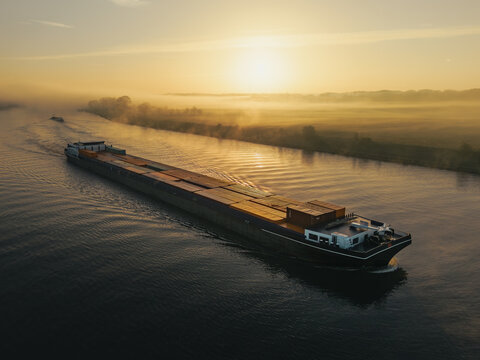 Aerial view of a large cargo barge navigating a calm river during a misty sunrise in Kessel, Limburg, Netherlands.