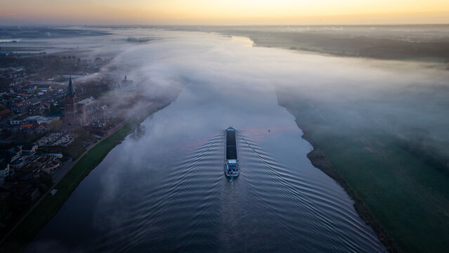 Aerial view of a cargo ship navigating the river through thick morning mist past the historic village of Kessel, Limburg, Netherlands.