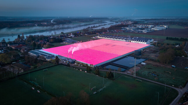 Aerial view of a large greenhouse illuminated with vibrant pink LED lights at dusk, situated near a river with surrounding fields and industrial buildings in Kessel, Limburg, Netherlands.