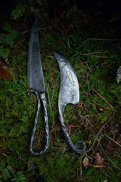 Long pointed knife and shorter curved blade rest side by side on forest moss in a close vertical photograph of textured metalwork with dark curled handles and natural greenery