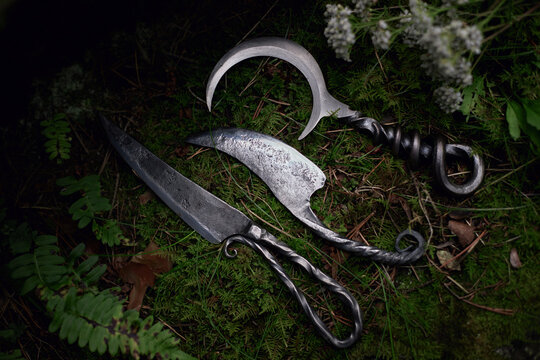 Three metal blades with twisted ornamental handles lie diagonally on forest moss beside small white flowers in a rustic still life photograph of traditional metalwork