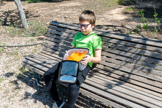 Child sits on a bench in a park and looks at a backpack while holding an orange object