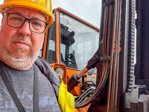 Industrial Worker in Hard Hat and Hi-Vis Vest Taking Selfie Next to Orange Forklift at Job Site
