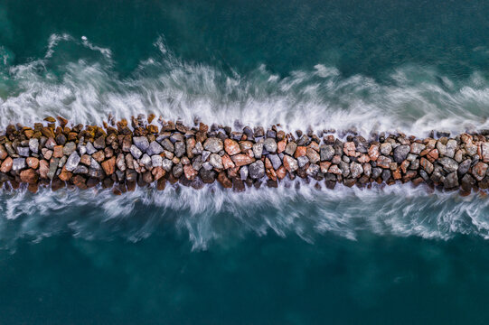 Aerial view of a stone jetty with waves crashing against the rocks in the turquoise ocean at Newport Beach, California, United States.