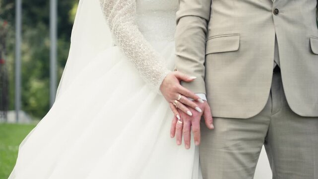 A bride and groom stand together in a garden, the bride holds a bouquet while the groom has a firm grip on her hand.