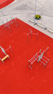 Aerial view of a modern outdoor fitness park with red rubber flooring, metal exercise equipment, and benches in Riga, Latvia.