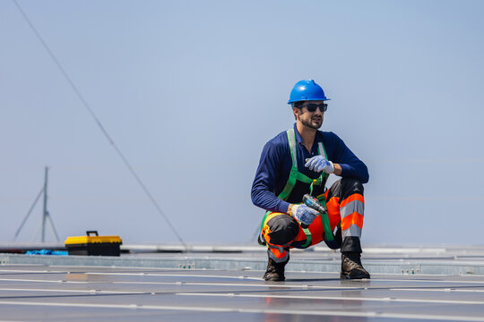 Solar technician in safety harness and hard hat holding tool while inspecting photovoltaic system, Engineer man working on renewable energy installation and maintenance at solar power plant