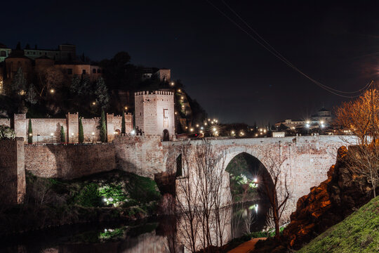 View of the illuminated Puente de San Martin stone bridge at night with medieval city walls and fortifications in Toledo, Castile-La Mancha, Spain.