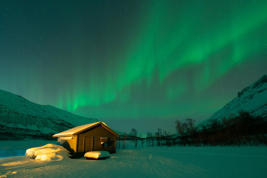 View of a small wooden cabin covered in snow under a vibrant display of green aurora borealis in the night sky in Norway.