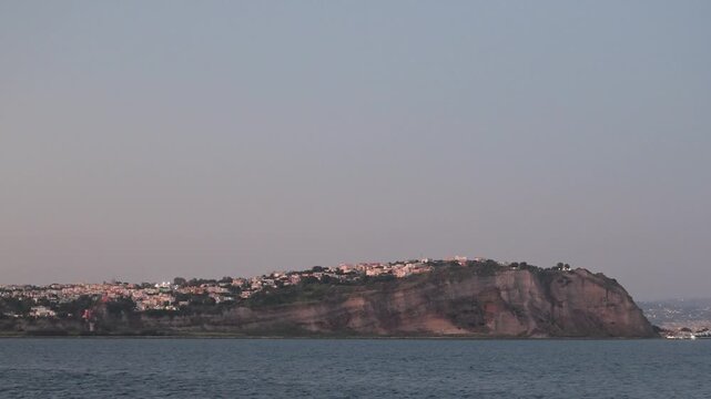 Monte di Procida&rsquo;s cliffside houses catch soft evening light above calm coastal water in a static wide shot.