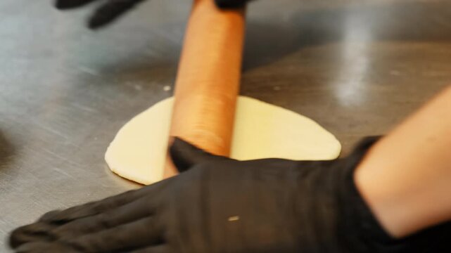Gloved baker rolls pastry dough across stainless worktable with wooden pin. Firm pressure flattens round piece preparing thin wrapper for filling