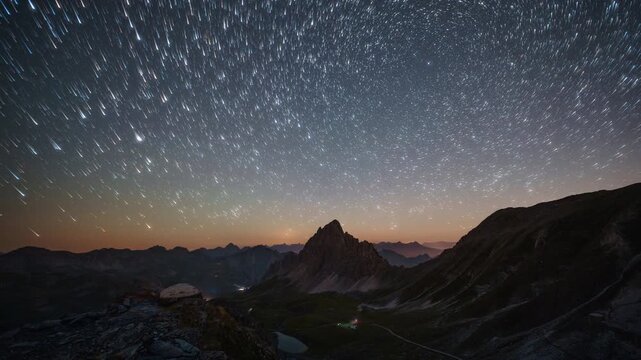 Long exposure timelapse of star trails swirling over a sharp mountain peak and faint valley lights on a clear night.