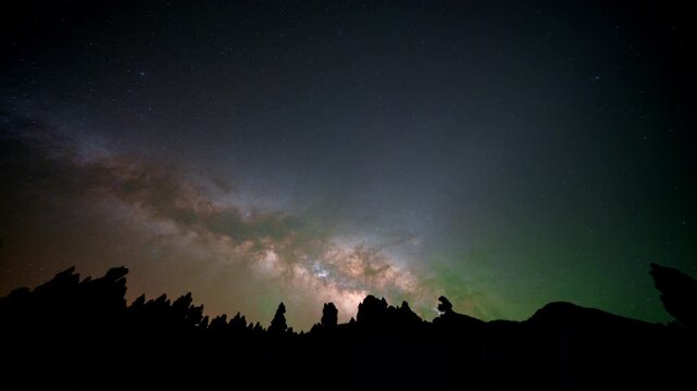 A glowing galaxy band rises across the sky above dark jagged rock formations with green airglow along the horizon.