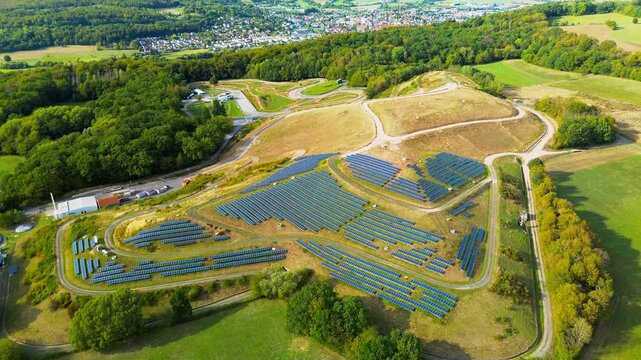 Aerial orbit above dense rows of solar panels covering a reclaimed landfill in the German countryside, with a works depot, sewage basins, forest and a town spread along the far ridge.