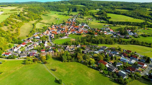 Aerial view directly over a German village, showing red roofs, a church tower, curving streets, private gardens and a small building site, surrounded by meadows, arable fields and forest.