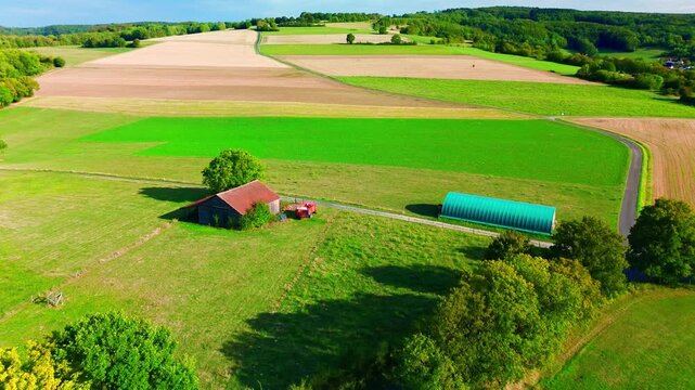 Aerial view of an old field barn with a red corrugated roof and a green storage tunnel standing beside a quiet country road, surrounded by a patchwork of stubble, worked soil and fresh crop.