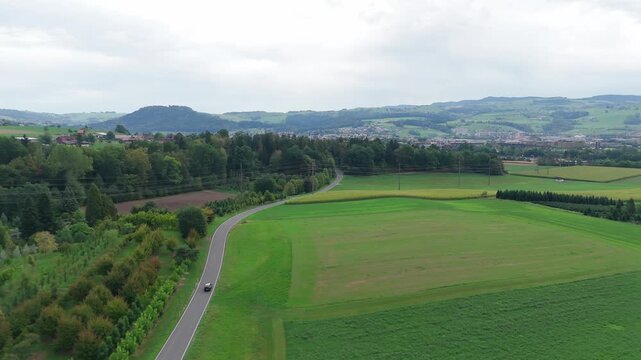 Drone view of a car driving along a winding country road through lush green fields, patches of forest, and rolling hills near Baar, Switzerland.