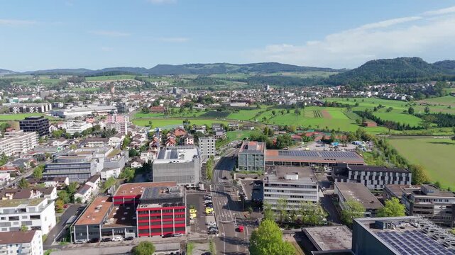 Drone view over the industrial and residential area of Baar in the Canton of Zug, Switzerland, showing modern commercial buildings, green fields, and hills under a sunny sky.