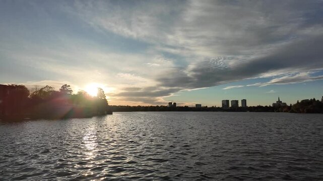 Scenic sunset over Herastrau Lake in Bucharest, Romania, with sunlight reflecting on the water beneath dramatic clouds and the distant city skyline.