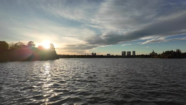 Scenic sunset over Herastrau Lake in Bucharest, Romania, with sunlight reflecting on the water beneath dramatic clouds and the distant city skyline.