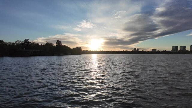 Scenic sunset over Herastrau Lake in Bucharest, Romania, with sunlight reflecting on the water beneath dramatic clouds and the distant city skyline.