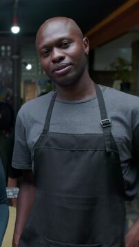 Confident African American male waiter wearing dark apron standing inside restaurant looking directly at camera with calm expression to project dedicated hospitality service