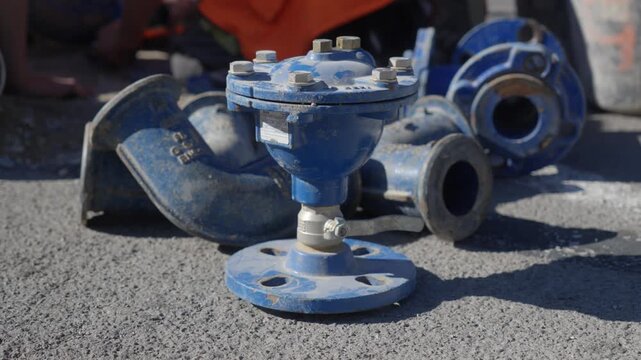 Camera slowly pushes toward blue flanged pipe fittings on sunlit asphalt