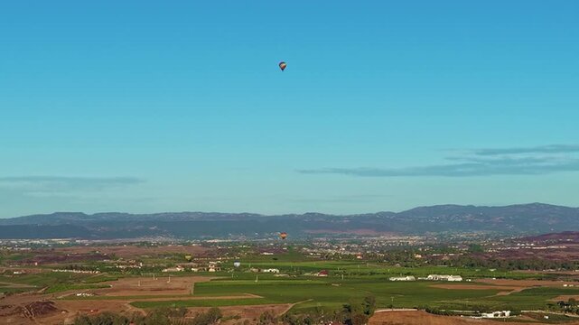 Hot air balloons drift above Temecula wine country farmland and distant hills, aerial orbit
