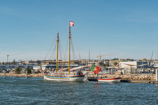 View of a traditional wooden sailing boat with a colorful hull and the Portuguese flag docked in the marina with buildings under a clear blue sky in Lagos, Faro District, Portugal.
